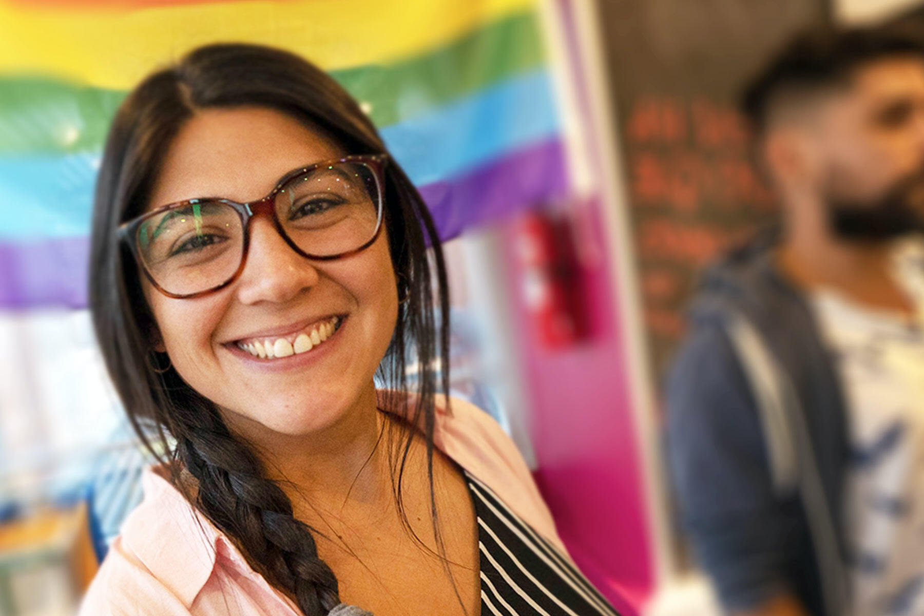 A young lady in one of our offices in Brazil, smiling warmly at you with a rainbow background blurred behind her, and a man with a beard standing, also blurred..
